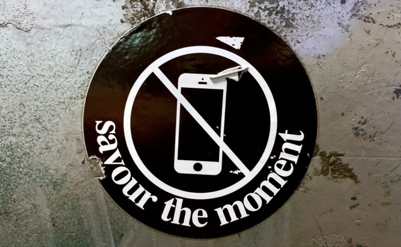 Sticker mit durchgestrichenem Smartphone und dem Text „Savour the moment“ auf rauer Betonwand – Symbol für digitale Achtsamkeit und Fokus.